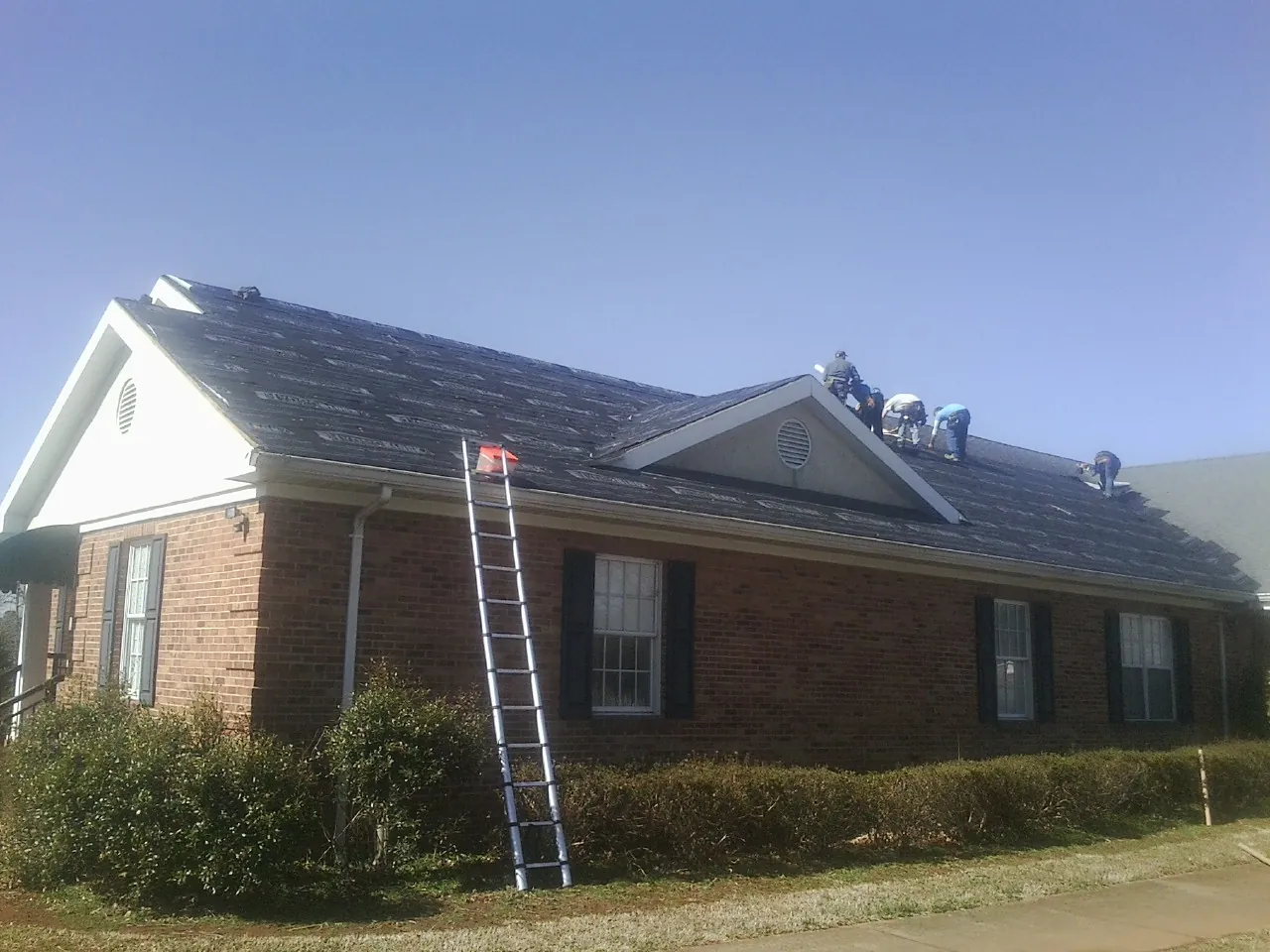 Crew tearing shingles off a brick ranch roof with a ladder set — Emergency Roof Repair work by our crew in Wagram