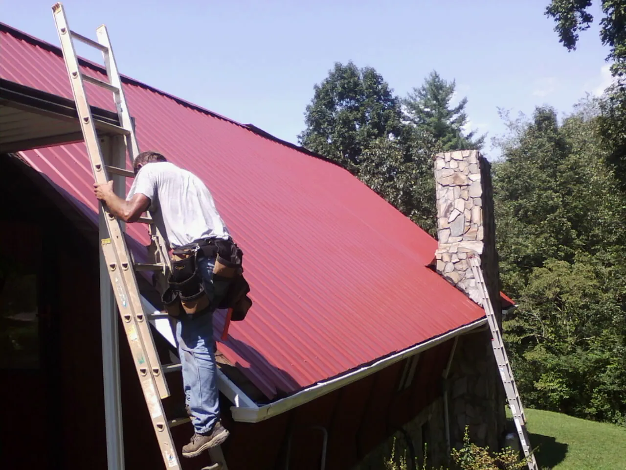 Roofer on a ladder beside a red metal roof and stone chimney — Soffit & Fascia Repair by Roofing Brothers in Wagram